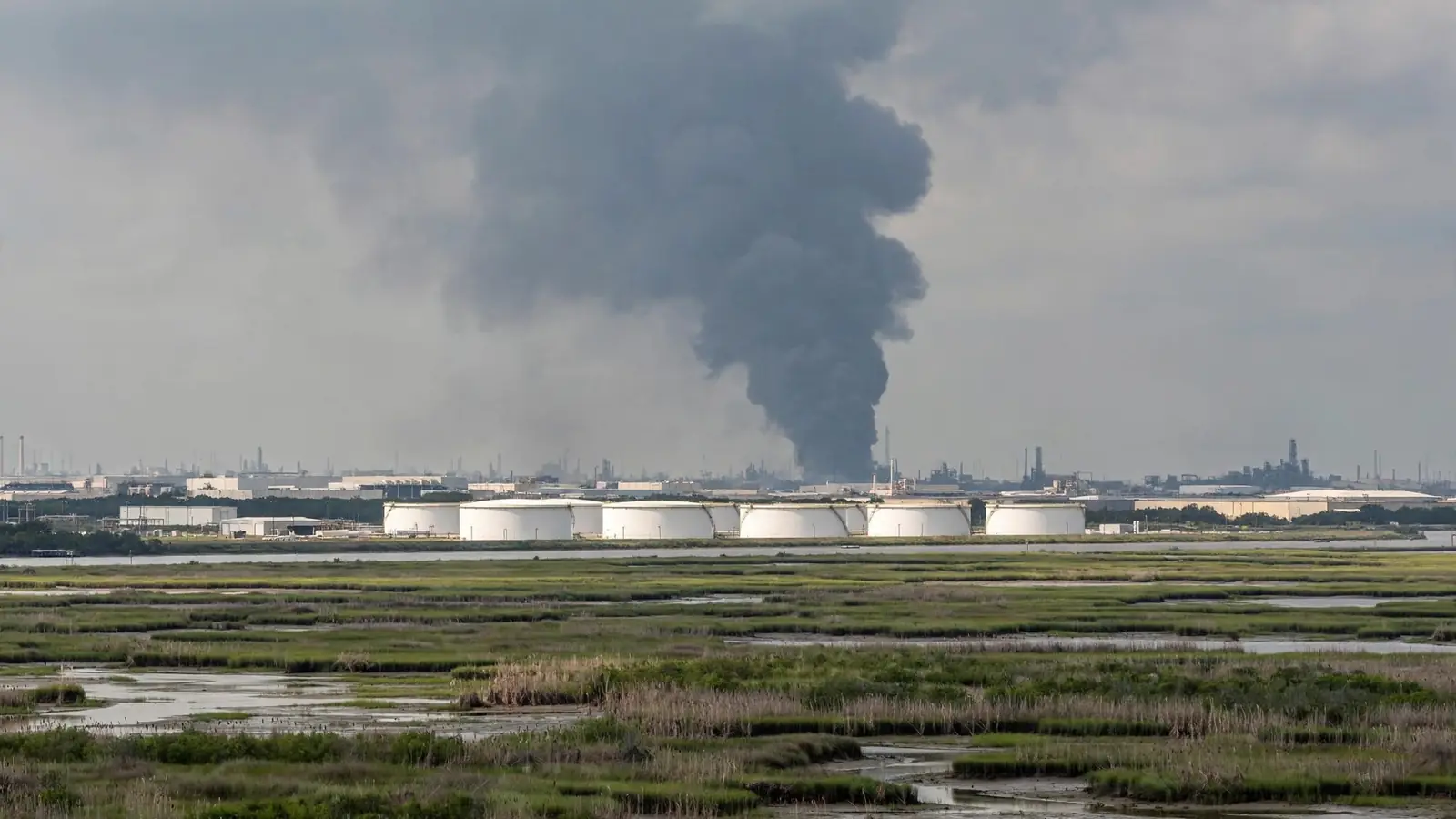 Dark smoke plume rising over industrial storage tanks seen from a distance